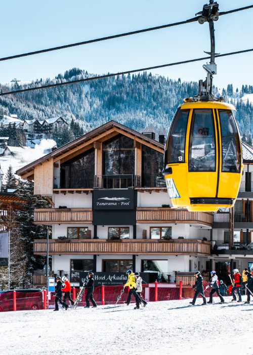 Cabinovia gialla a Corvara sopra le piste con sciatori davanti all’Hotel Arkadia