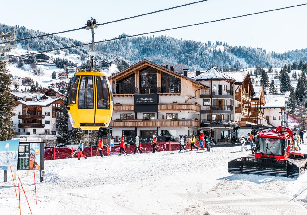 Cabinovia gialla sopra le piste da sci davanti all’Hotel Arkadia con sciatori e gatto delle nevi