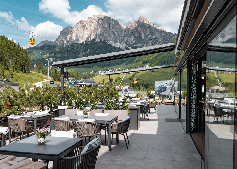 Terrazza del bistrot con tavoli e sedie nere, piante, incorniciato dalle maestose Dolomiti